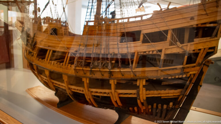 Close view of wooden ship model showing exposed ribs, hull planking, deck beams, internal framing, and rigging lines.