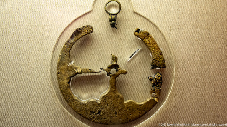 Corroded circular iron fitting with broken ring, projecting hardware, and small fragments displayed in a museum case.