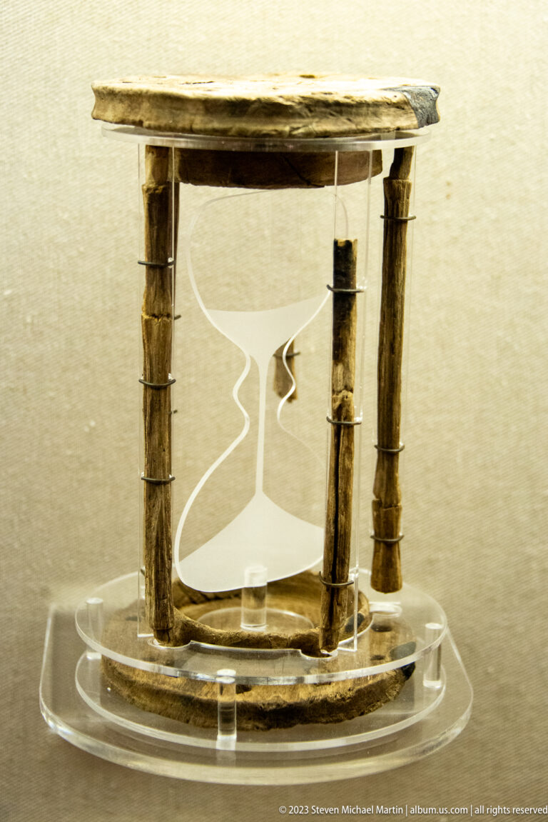 Small weathered timber framework with upright posts and curved wooden base displayed on a clear museum mount.