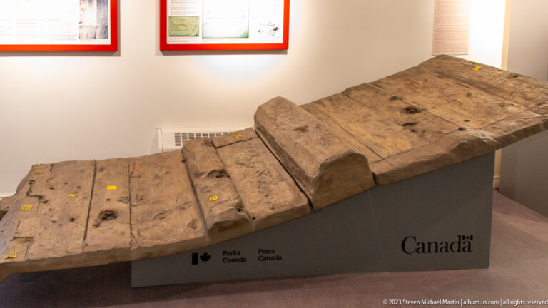 Large weathered ship timber with stepped profile, holes, and numbered feature markers displayed on a Parks Canada plinth.
