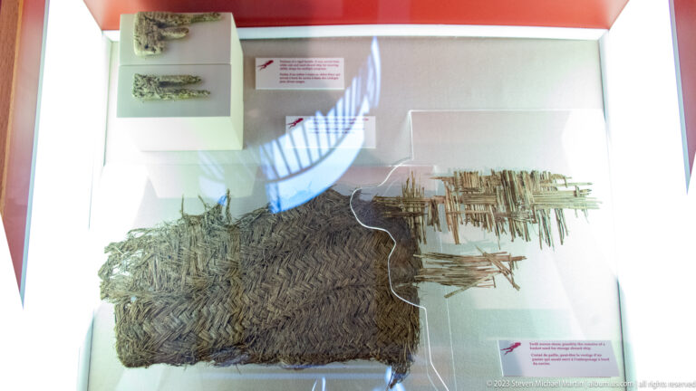 Dark woven fiber mat and loose reed-like fragments suspended in a museum display case.