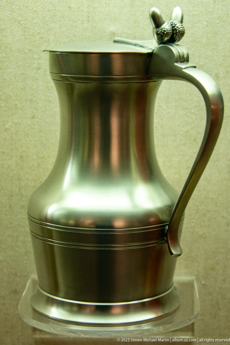 Rounded pewter vessel with curved handle, hinged lid, low foot, and dark metallic surface.