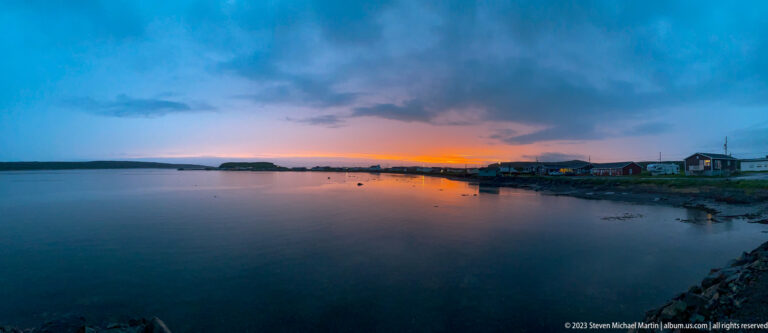 Wide sheltered inlet under blue clouds and an orange horizon, with low shoreline buildings at right.