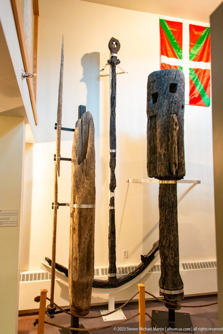 Large weathered wooden and iron implements displayed upright beneath a red, green, and white Basque flag.