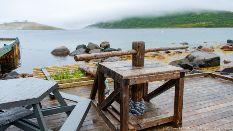 Weathered wooden work table on a harbor platform beside rocks, floating structures, and a foggy green headland.