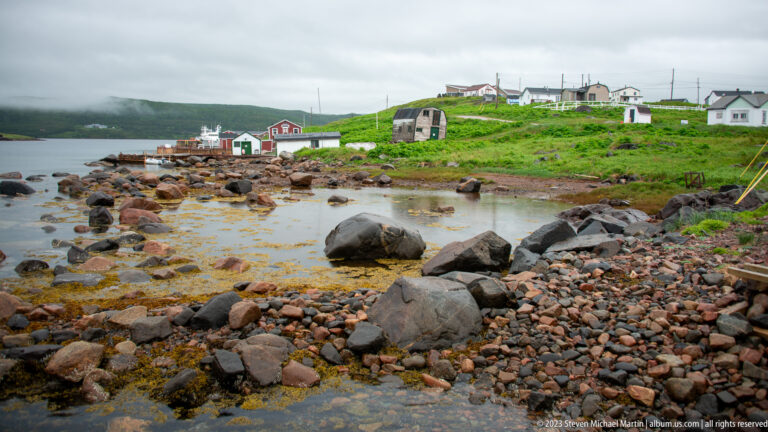 Rocky, seaweed-covered tidal cove with red-and-white waterfront buildings and houses on a green slope.