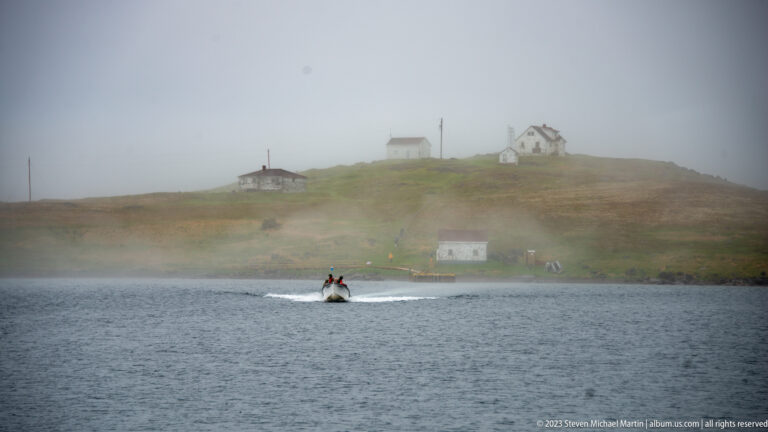 Small motorboat crossing gray harbor water below scattered hillside houses partly obscured by fog.