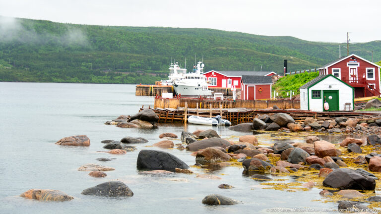 Red Bay waterfront with red-and-white buildings, timber wharf, moored white vessel, rocks, and seaweed.