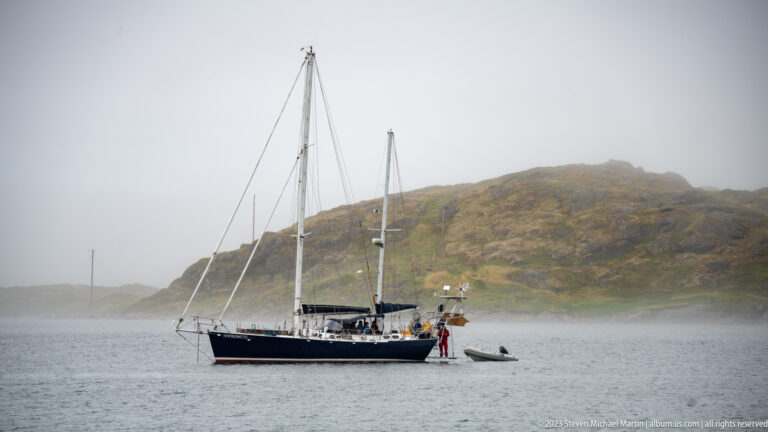 Dark-hulled two-masted sailing yacht at anchor with a small inflatable tender astern and foggy rocky hillside behind.