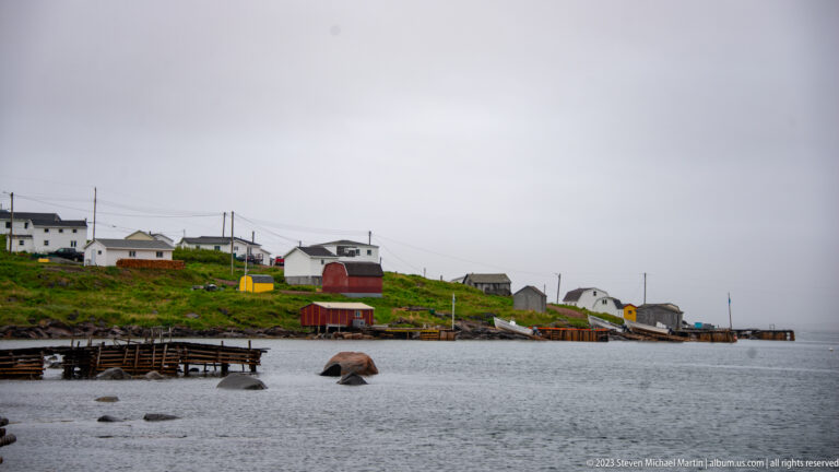 Scattered houses, sheds, wharves, and utility lines along a low green harbor shore under gray skies.