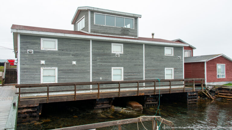 Weathered gray waterfront building on timber supports with white-trimmed windows, raised upper section, and wooden walkway.