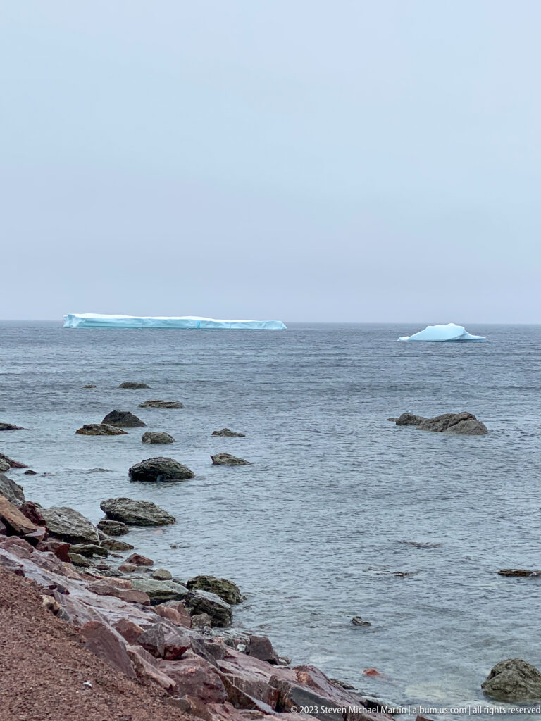 Two low blue-white icebergs offshore beyond scattered dark rocks and a reddish coastal edge.