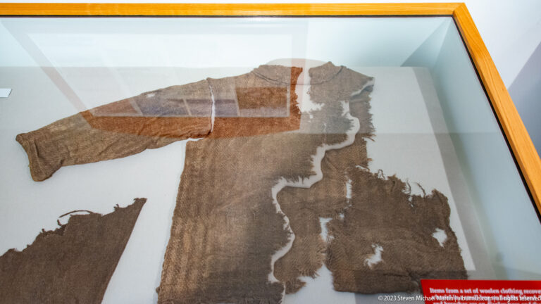 Large brown woven textile fragments with seams and missing areas displayed flat in a museum case.