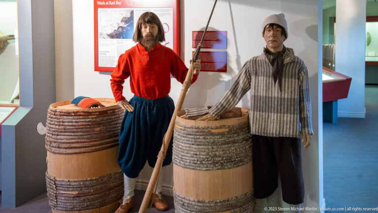 Two Basque whaler mannequins beside large wooden barrels, one in red holding a long pole and one in striped clothing.
