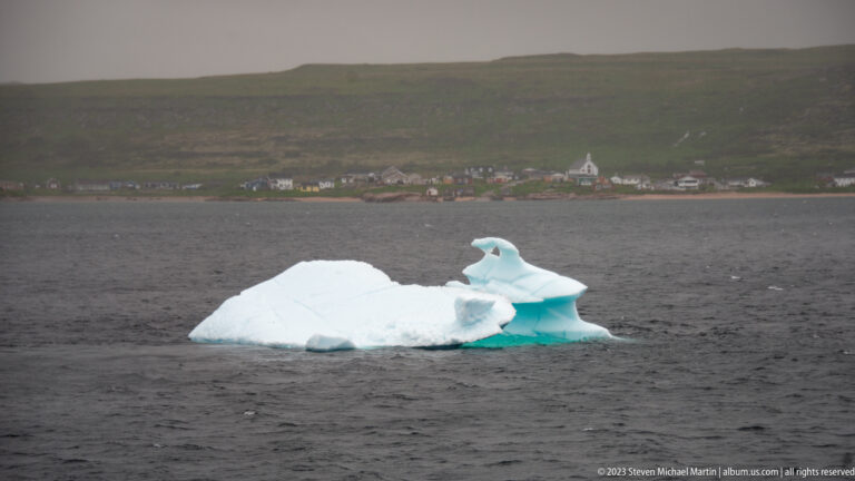 Low white iceberg with turquoise edges floating before a coastal village and green hillside.