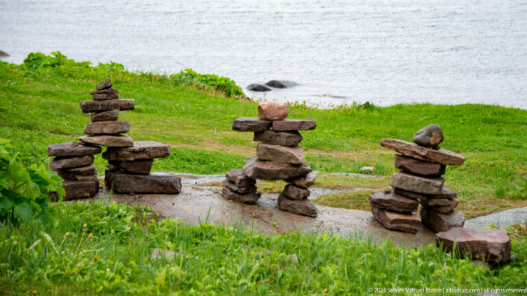 Several small stacked-stone humanlike figures on a grassy shore beside gray water.