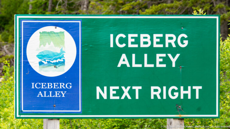 Green roadside sign reading 'ICEBERG ALLEY — NEXT RIGHT' beside a blue iceberg emblem.