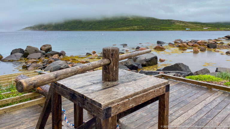 Heavy wooden platform with central post and long horizontal beam on a deck beside a rocky, foggy shoreline.