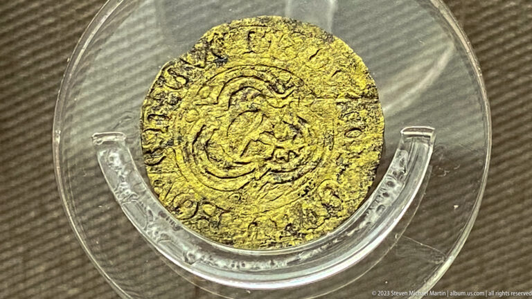 Small worn gold-colored coin on a clear mount with central emblem and circular lettering.