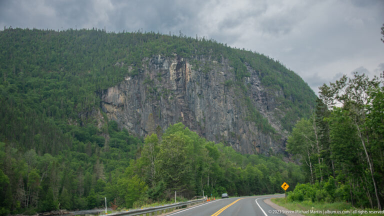 Two-lane road curving below a tall exposed rock cliff surrounded by dense forest.