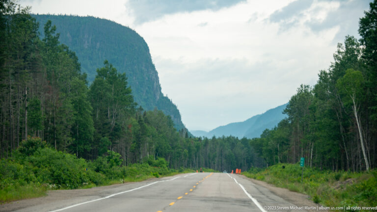 Straight two-lane highway leading between forested mountains beneath a cloudy sky.