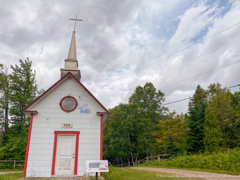 Small white wooden chapel dated 1911 with red trim, oval opening, steeple, and cross.