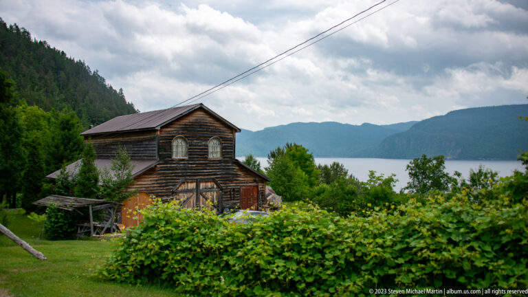 Weathered dark timber building among shrubs with the fjord and forested mountains behind it.