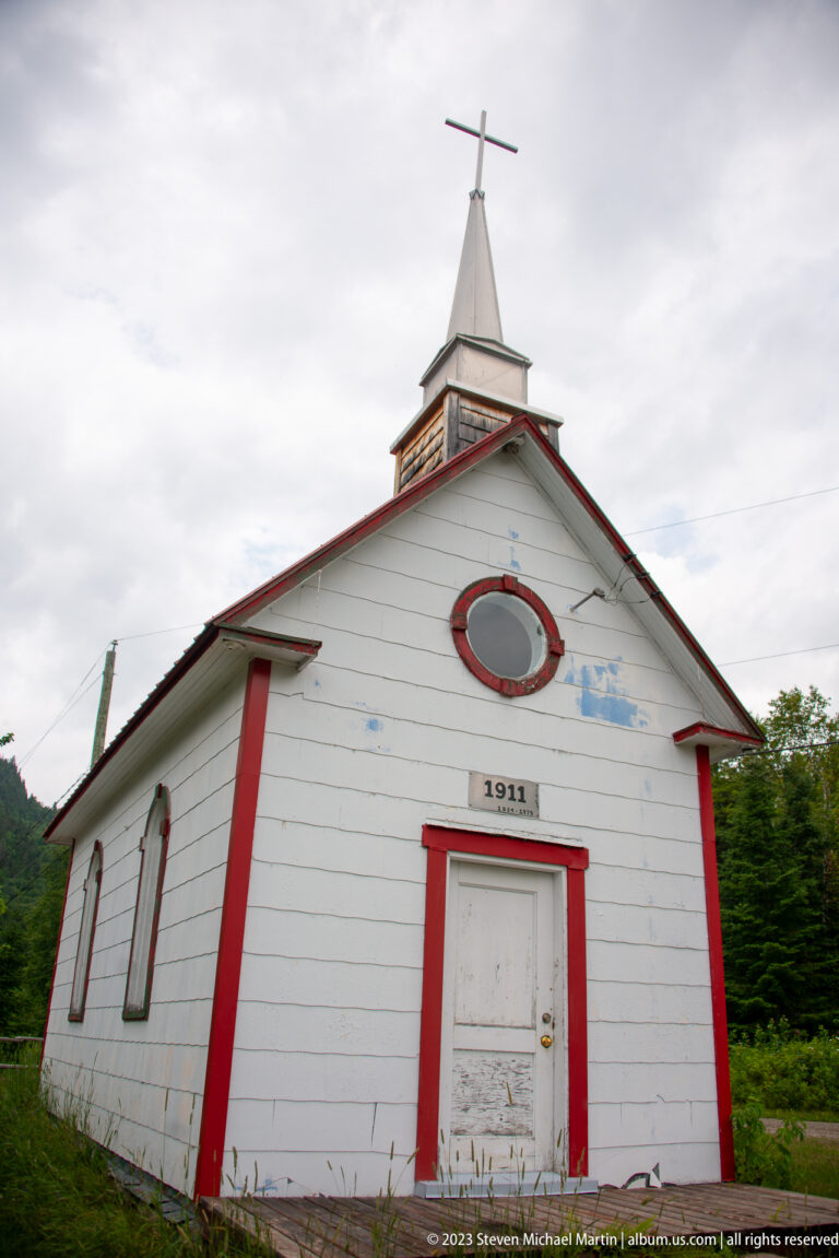 Weathered white 1911 chapel with red trim, narrow windows, steeple, and cross viewed from the front corner.
