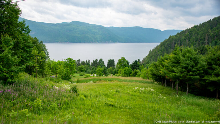 Green meadow and trees descending toward broad fjord water backed by forested mountain ridges.