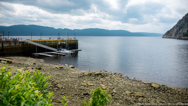 Floating dock and gangway beside a rocky shore on calm fjord water with mountains beyond.