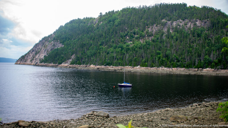 Small sailboat anchored in a rocky cove below a steep forested cliff on the Saguenay Fjord.