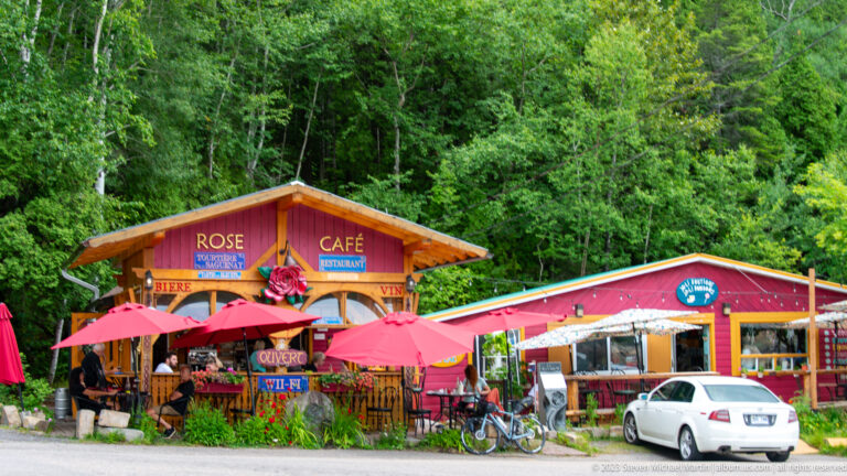 Brightly painted Rose Café with red umbrellas, bicycles, and a white car beside a forested road.