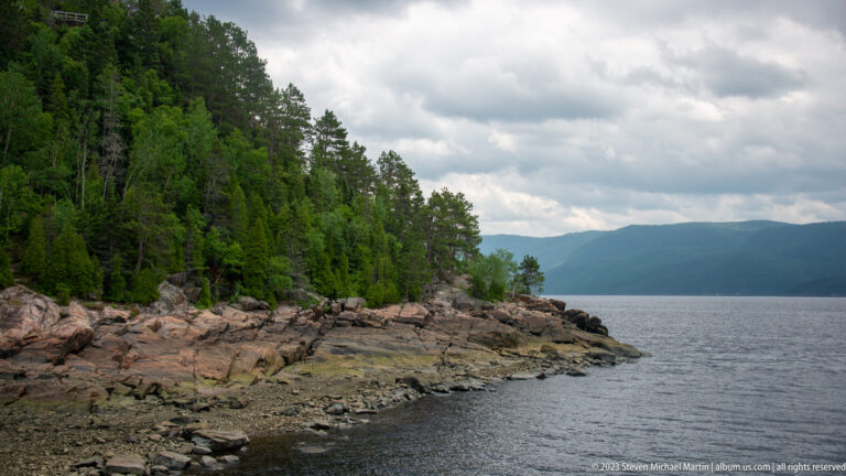 Jagged rocky point with dense trees extending into the Saguenay Fjord beneath cloudy skies.