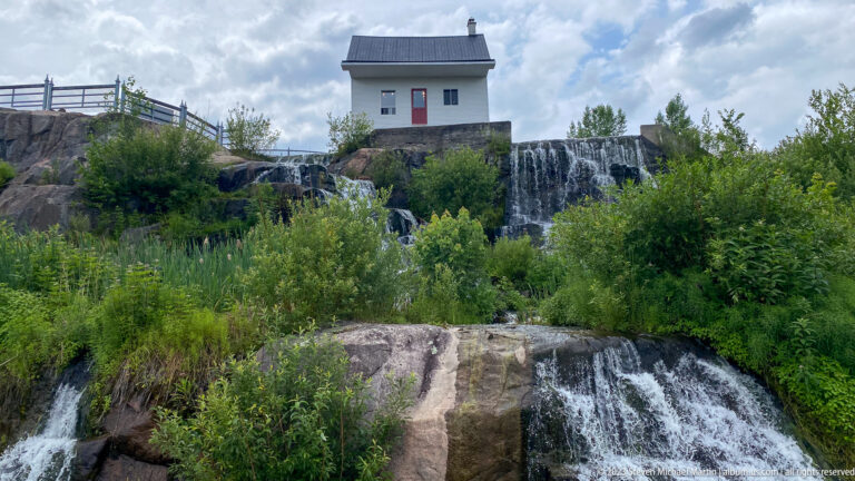 White utility building above multiple cascades flowing over broad rock ledges and dense greenery.