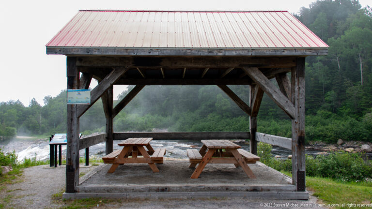 Open timber picnic shelter with two tables beside a forested river and visible rapids.