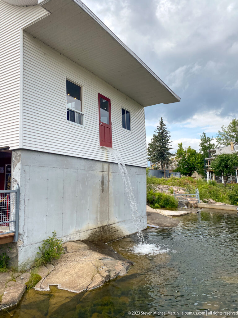 White industrial building with red doors beside a narrow controlled water channel and exposed bedrock.