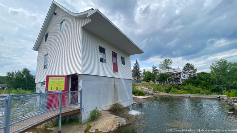 White utility building beside a narrow river channel, walkway railings, and a posted warning sign.
