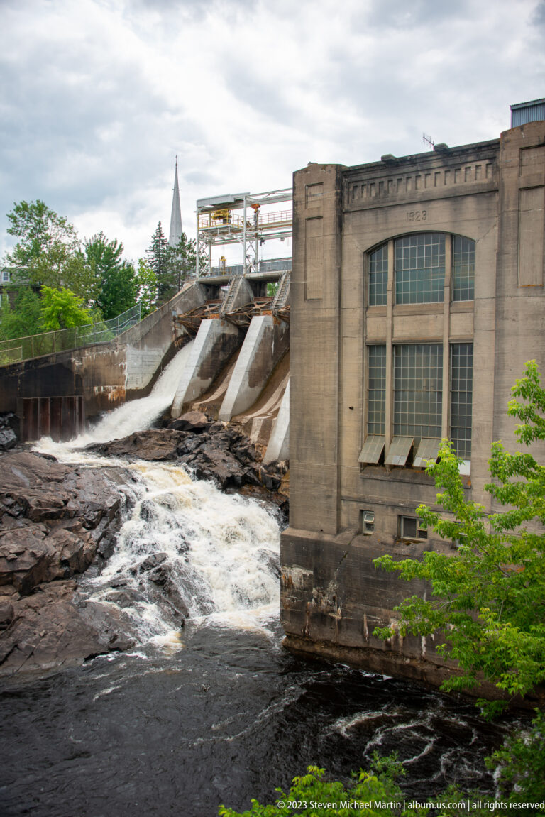 Concrete spillway releasing water beside a tall masonry hydroelectric powerhouse with large multi-pane windows.