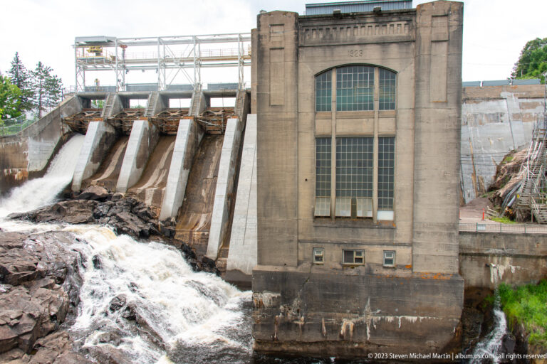 Barrage Chicoutimi powerhouse dated 1923 beside concrete spillways releasing water over bedrock.