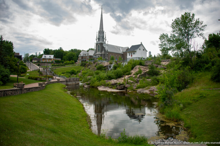 Gray stone Sacré-Cœur church with tall spire reflected in calm river water beyond landscaped lawns and rock.