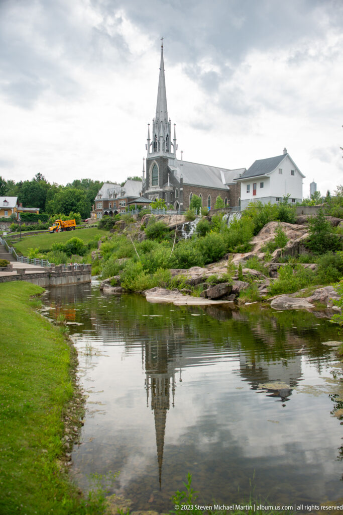 Sacré-Cœur church and tall spire reflected almost fully in calm river water with rocky landscaped banks.