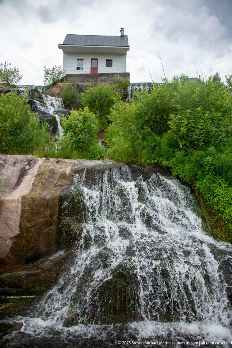 Broad cascade flowing over rounded bedrock with shrubs and a small white utility building above.