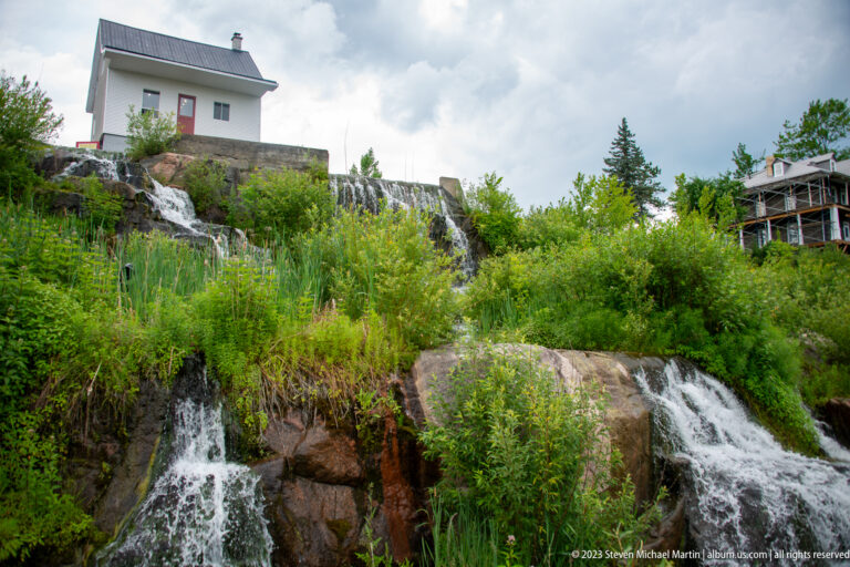 Multiple small waterfalls descending through dense green vegetation over brown bedrock below dam structures.