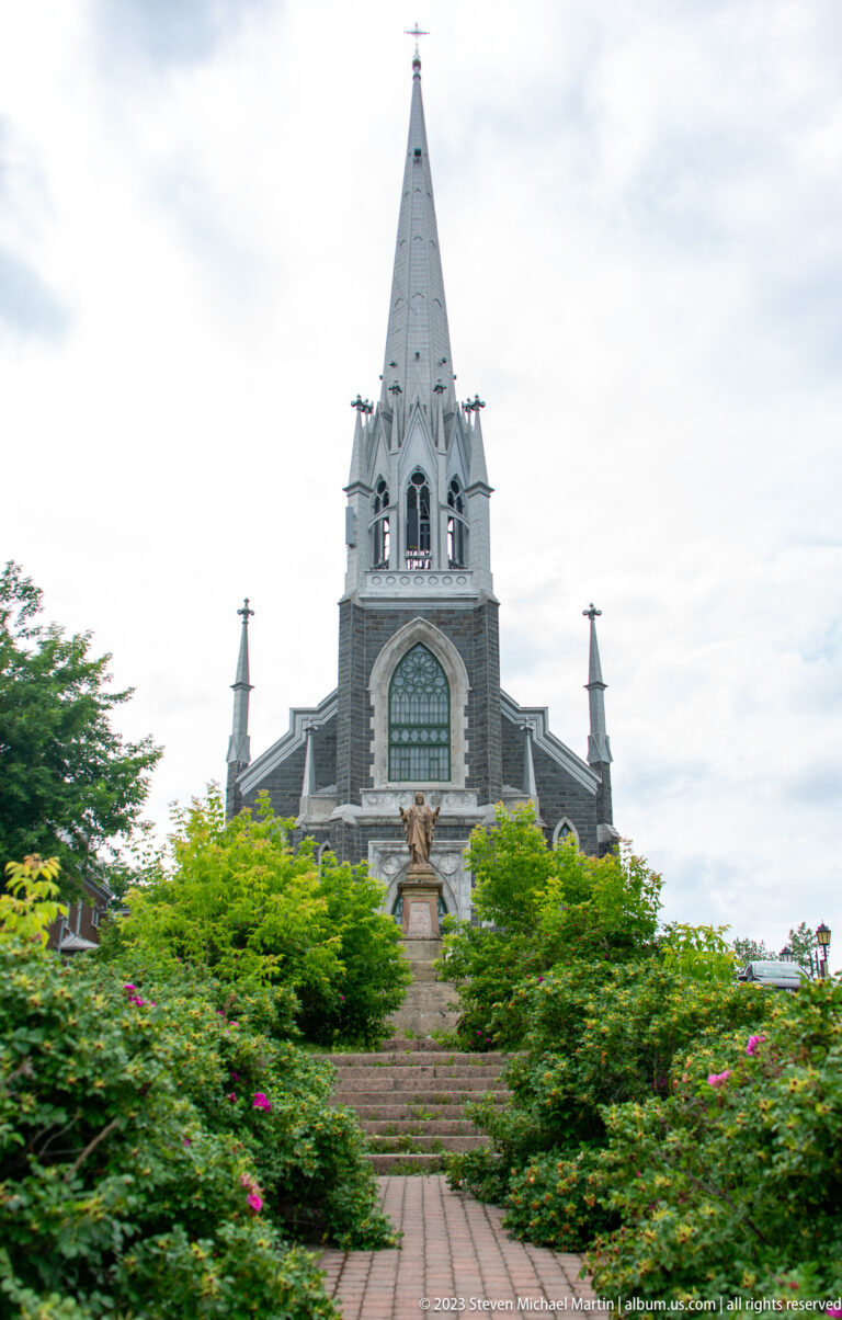 Neo-Gothic Sacré-Cœur church with dark stone façade, white spire, stairs, and flower garden.