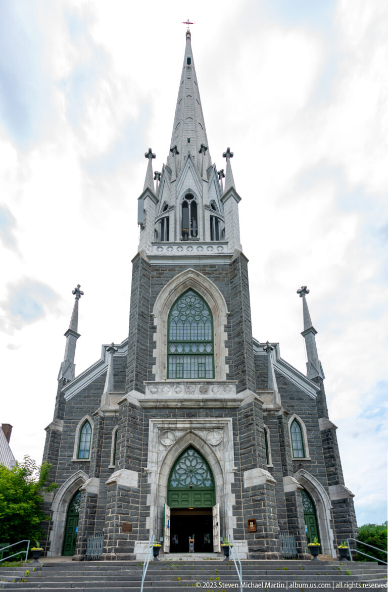 Frontal view of Sacré-Cœur church with pointed portal, large stained-glass window, pinnacles, and tall spire.