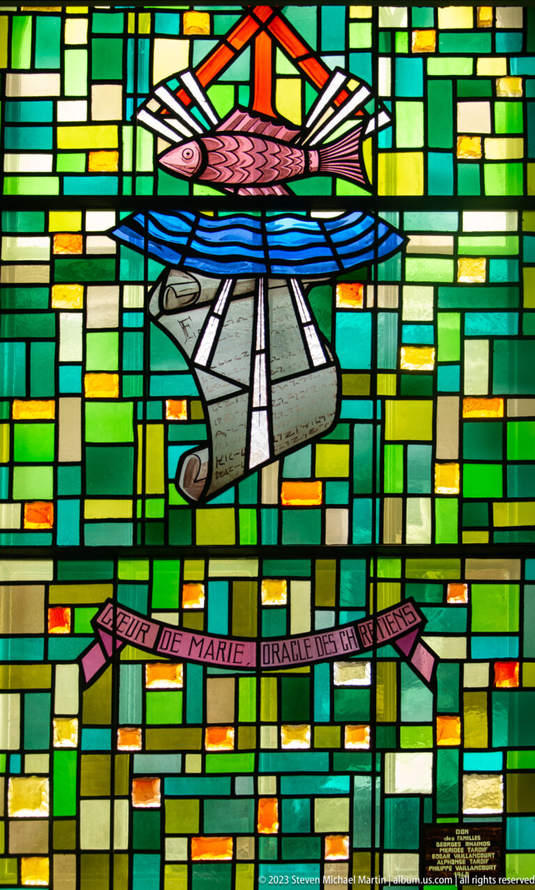 Colorful stained glass with stylized boat, small church, and pink banner bearing a Marian inscription.
