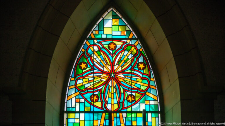 Pointed-arch stained-glass window with colorful radial flower-like pattern in red, blue, yellow, green, and white.