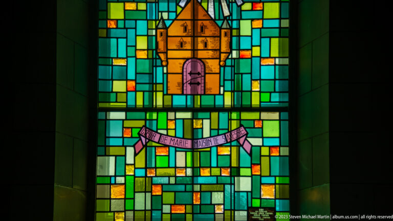 Stained-glass panel with orange chapel above a pink Marian banner, surrounded by green and blue geometric glass.