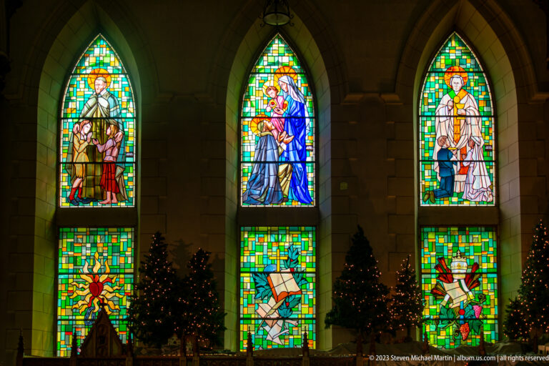 Three tall stained-glass windows with religious figures above smaller symbolic panels and decorated Christmas trees below.