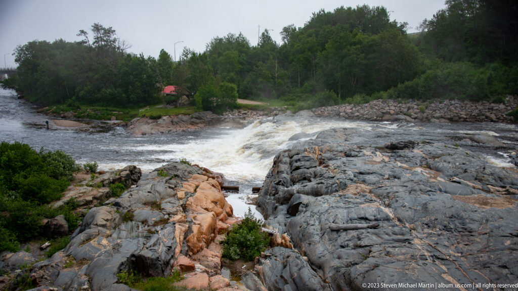 White-water rapids flowing through orange and gray bedrock with forest and a small red-roofed shelter beyond.